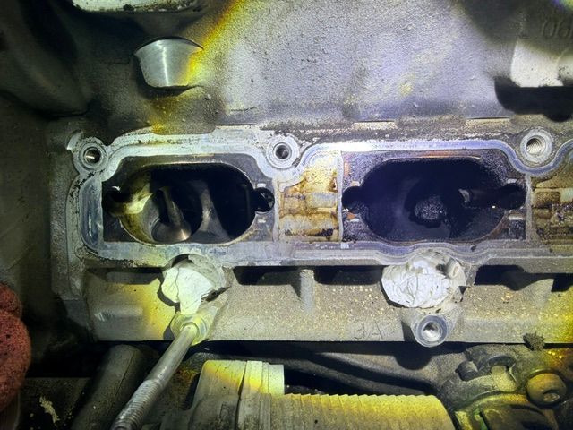Close-up of a dirty engine intake manifold with two open ports and a hand holding a tool.