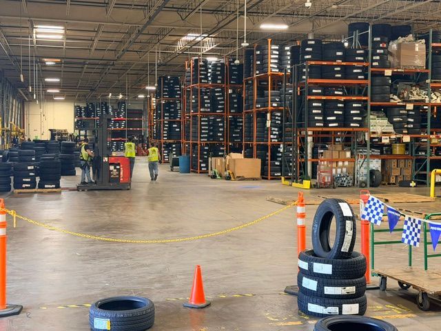 Warehouse interior with stacked pallets, orange cones, tire obstacles, and people working in the distance