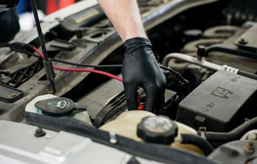 Black-gloved hand checking car battery with jumper cables in an engine bay