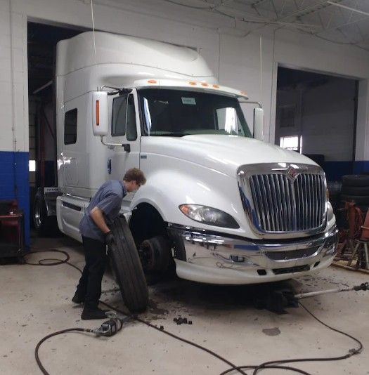 White semi-truck in a garage, with a mechanic changing a front tire.
