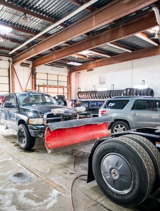 Two trucks in a garage with a red snowplow blade in front and a tire in the foreground