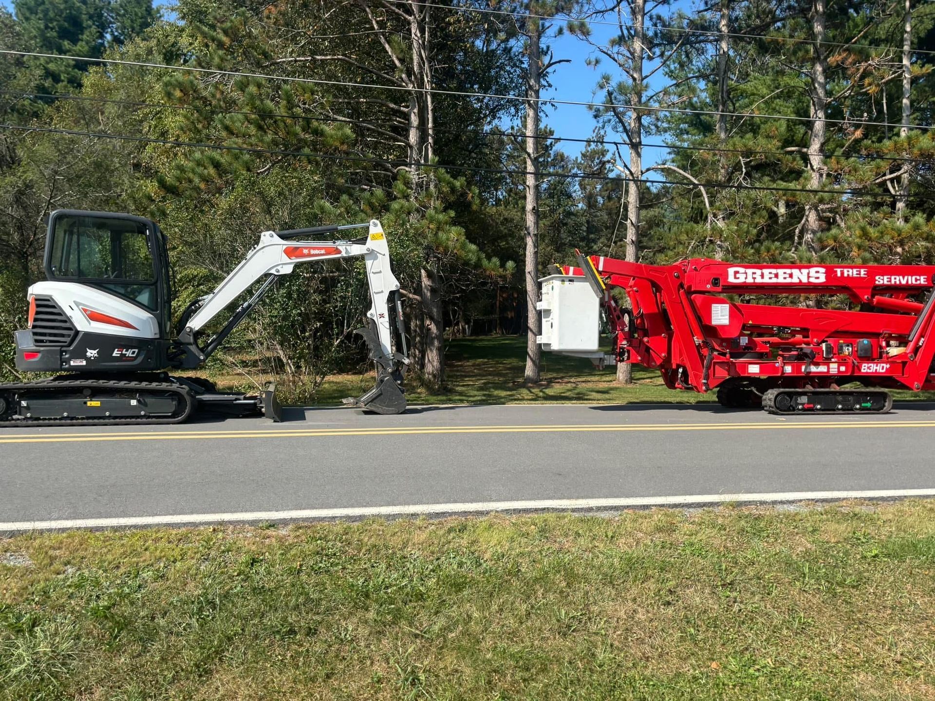 Excavator and red industrial machine on roadside near trees.
