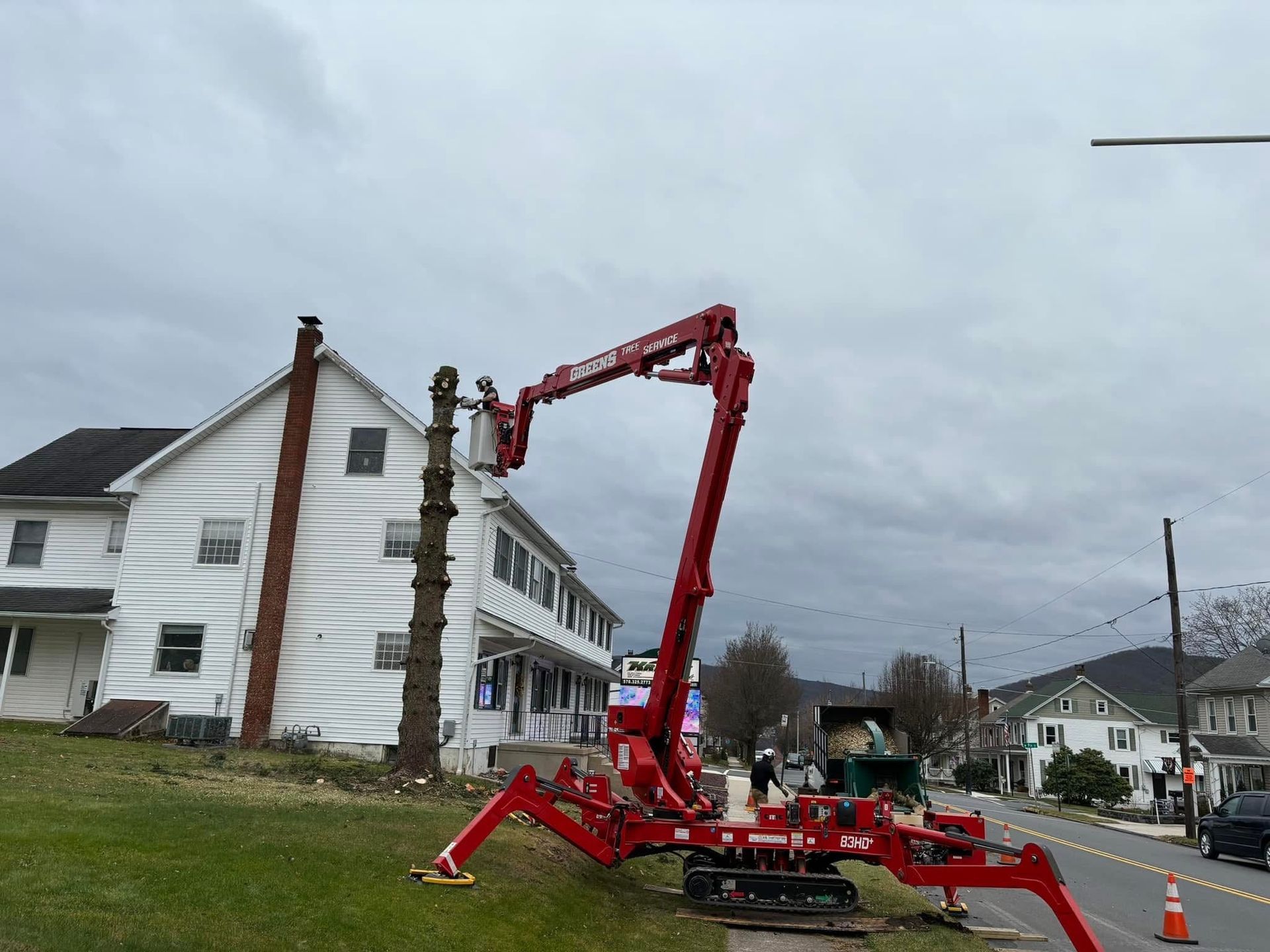A red spider lift trimming a tree in front of a white two-story house, overcast sky.