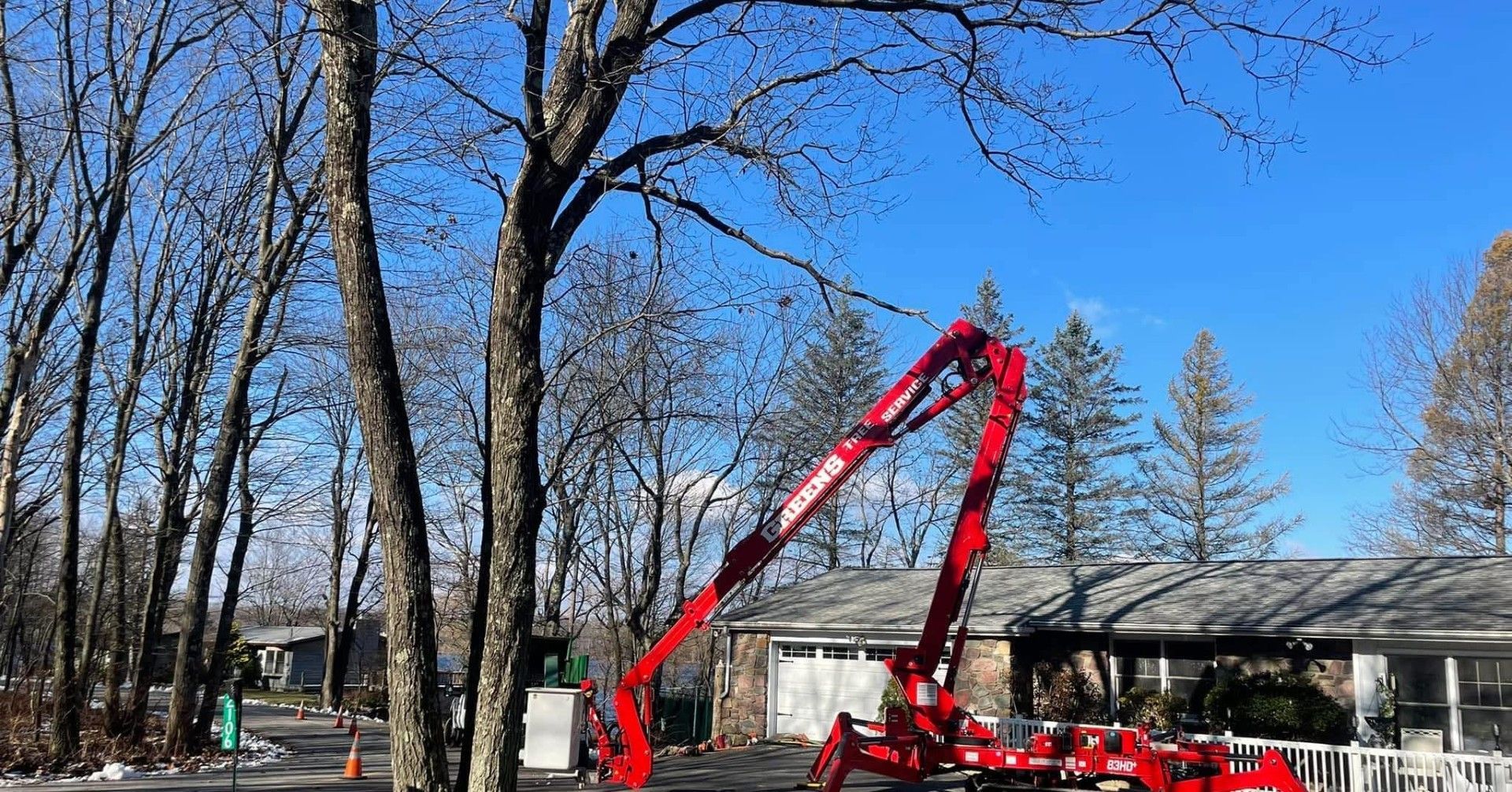 Red tree trimming lift reaching toward leafless tree in front of a house. Bright blue sky.