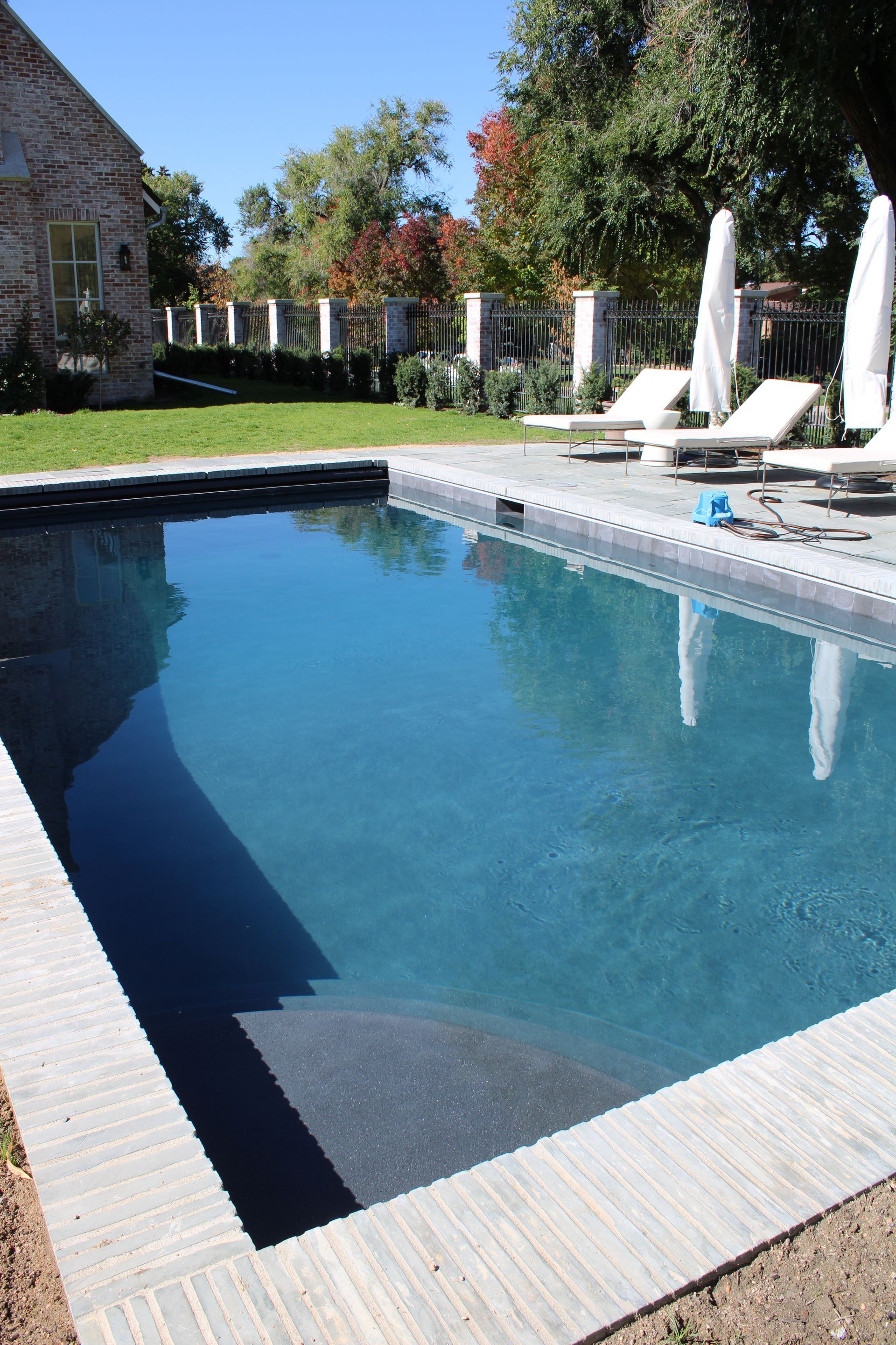 Rectangular pool with blue water. Beige tiled border. Loungers and umbrellas nearby. Building in the background.