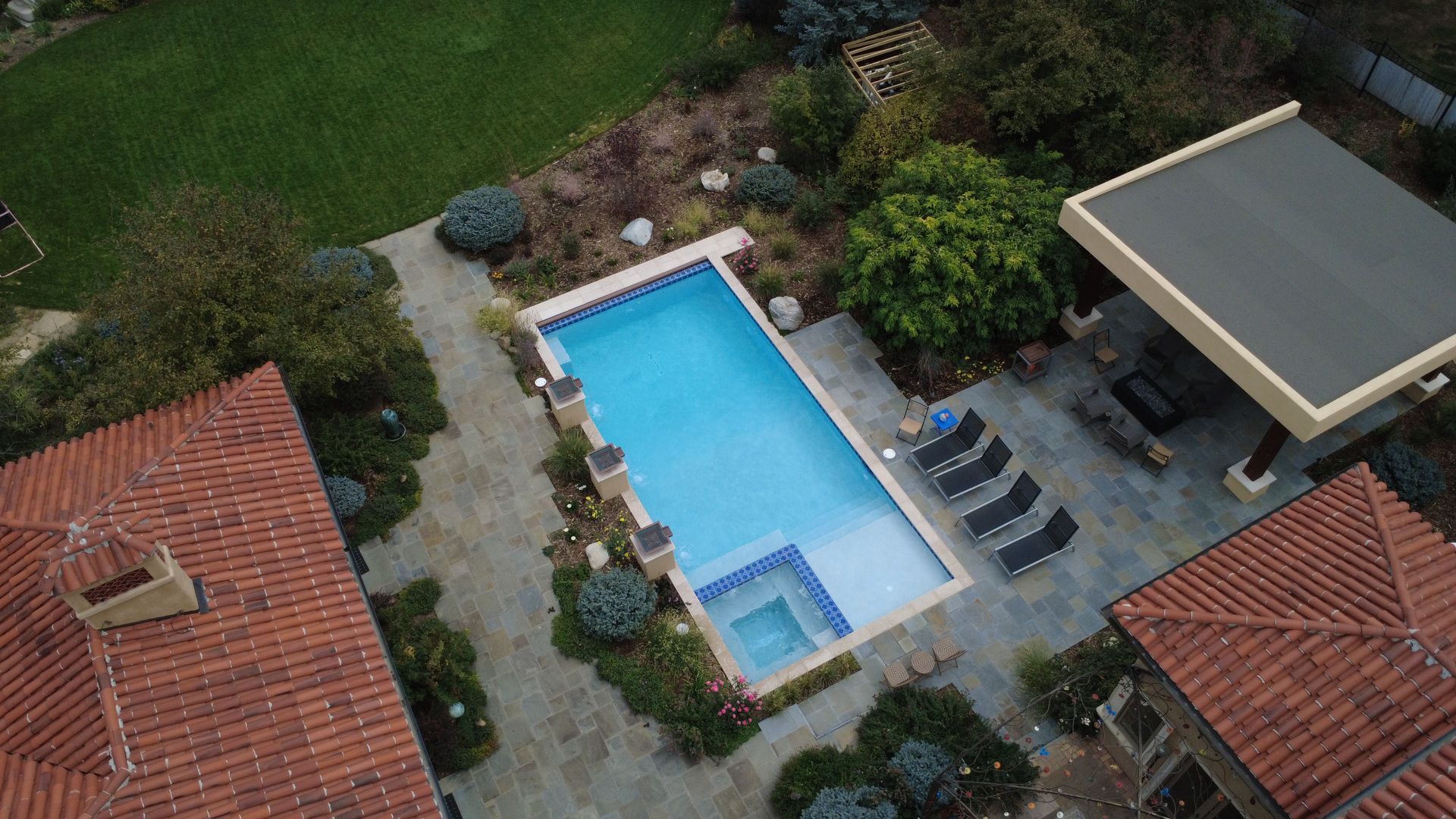 Aerial view of a rectangular pool with a spa, surrounded by a patio and landscaping, between two buildings with terracotta roofs.