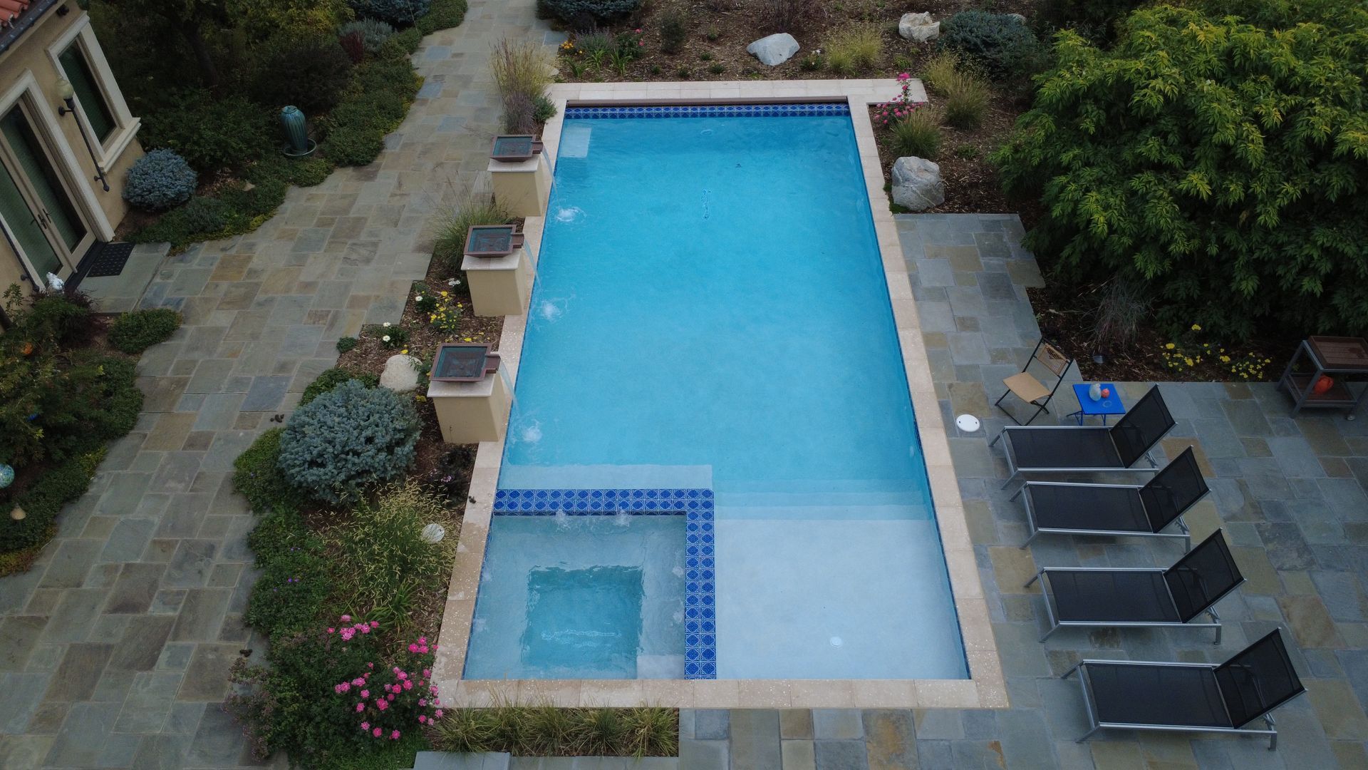 An overhead shot of a rectangular pool with a built-in spa, surrounded by greenery and patio.