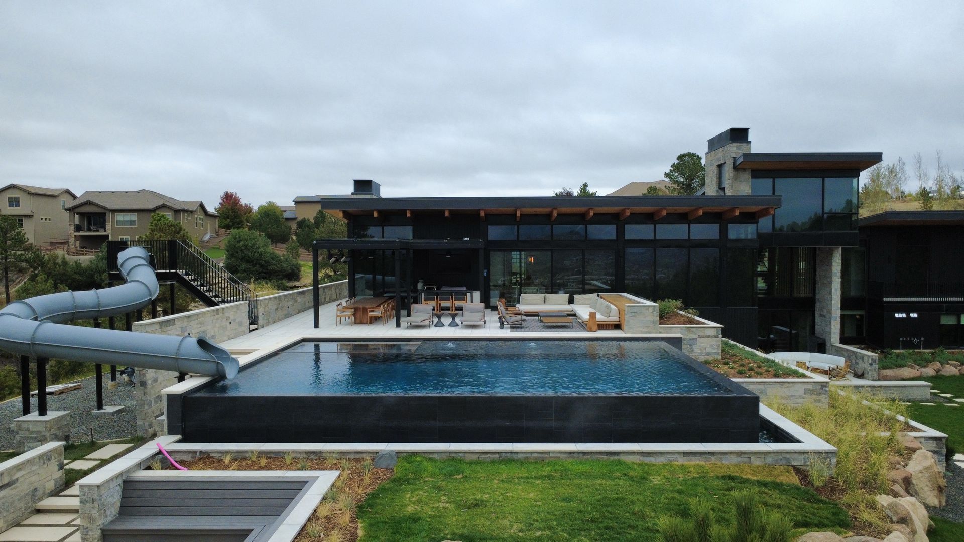 Modern house with pool and water slide on a hillside under a cloudy sky.