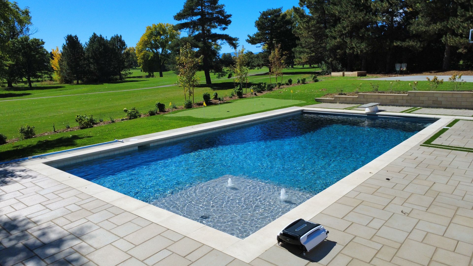 Rectangular swimming pool with blue water on a sunny day. Pavers surround the pool, and grass and trees are in the background.