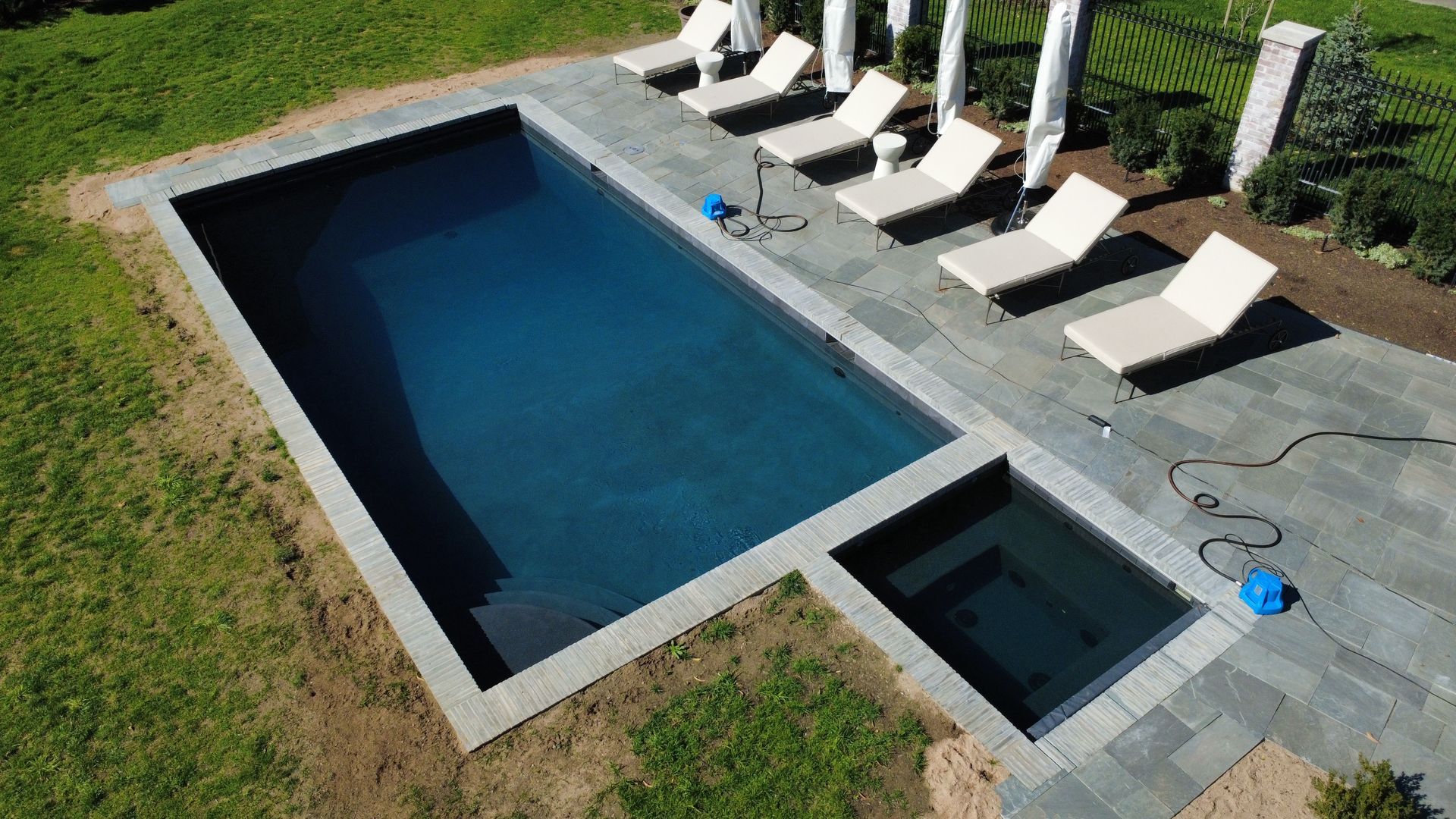 Aerial view of a rectangular pool with a connected jacuzzi, surrounded by lounge chairs and paving stones on a lawn.