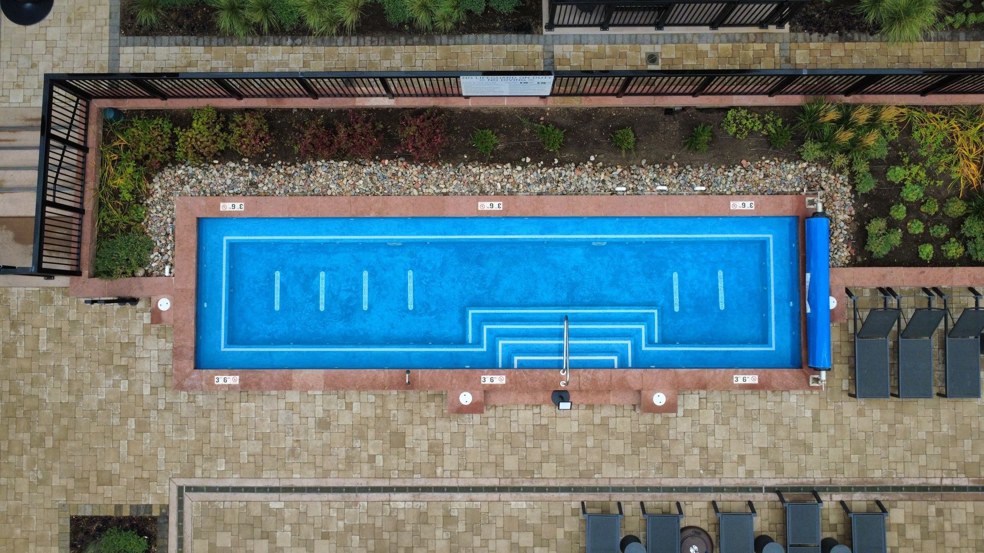 Overhead view of a rectangular blue swimming pool with steps, surrounded by stone paving, plants, and black lounge chairs.