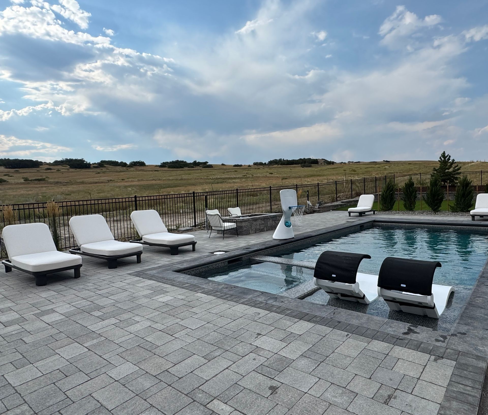 Outdoor pool with lounge chairs and grills on a patio, with a field and cloudy sky in the background.