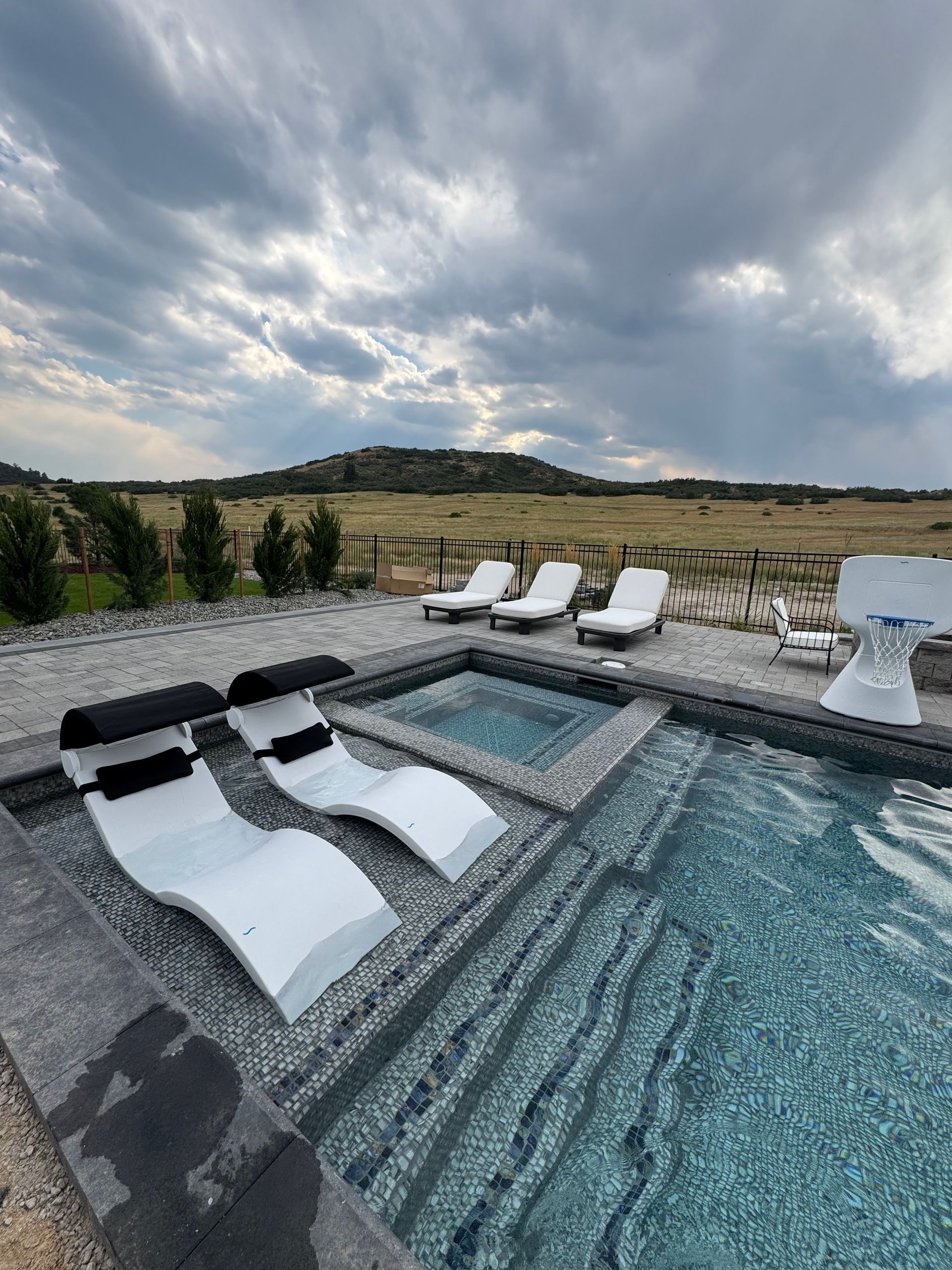 Pool with white lounge chairs, spa, and scenic landscape under cloudy sky.