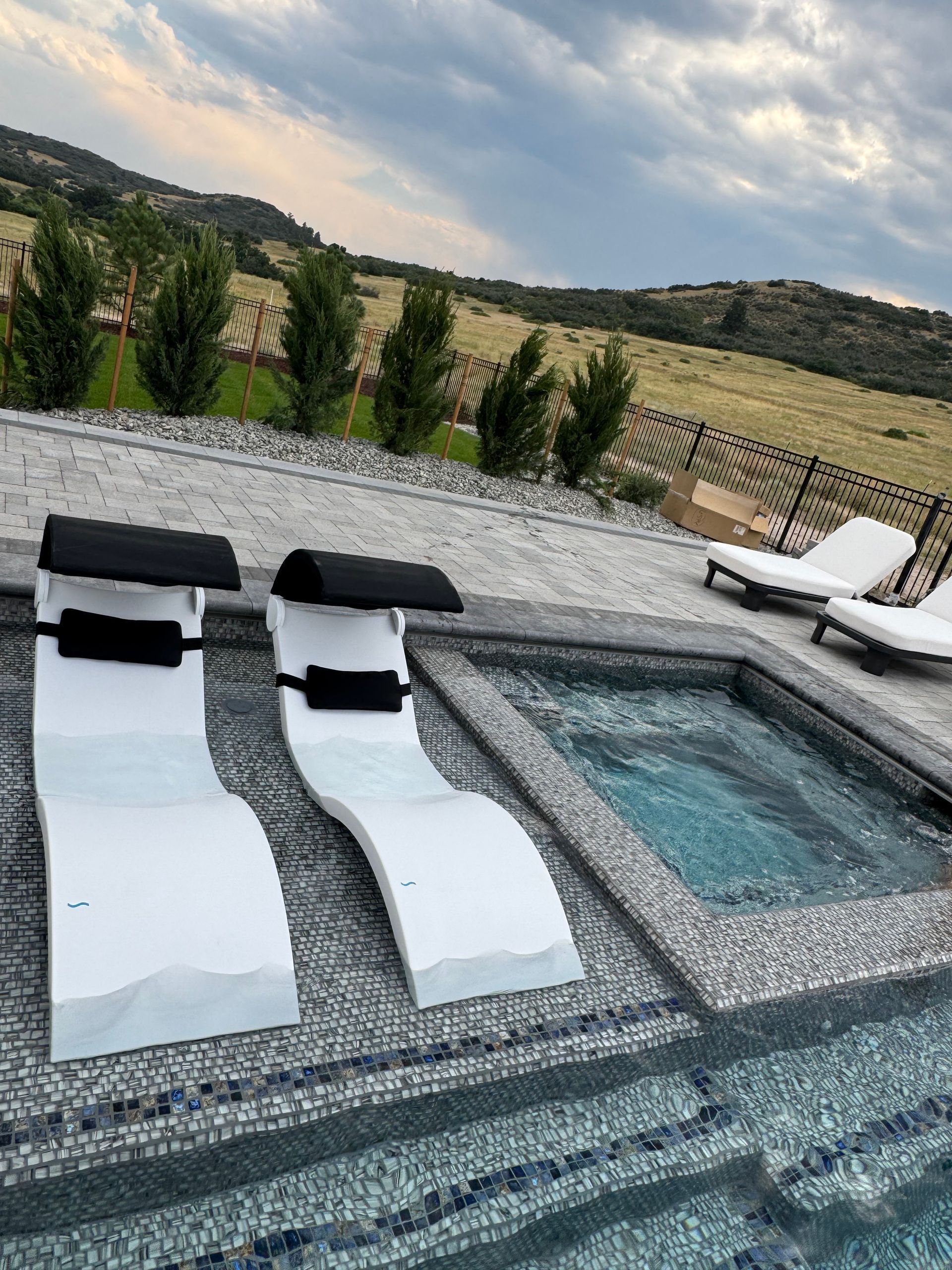 Two white chaise lounges by a pool and hot tub with a mountain backdrop.