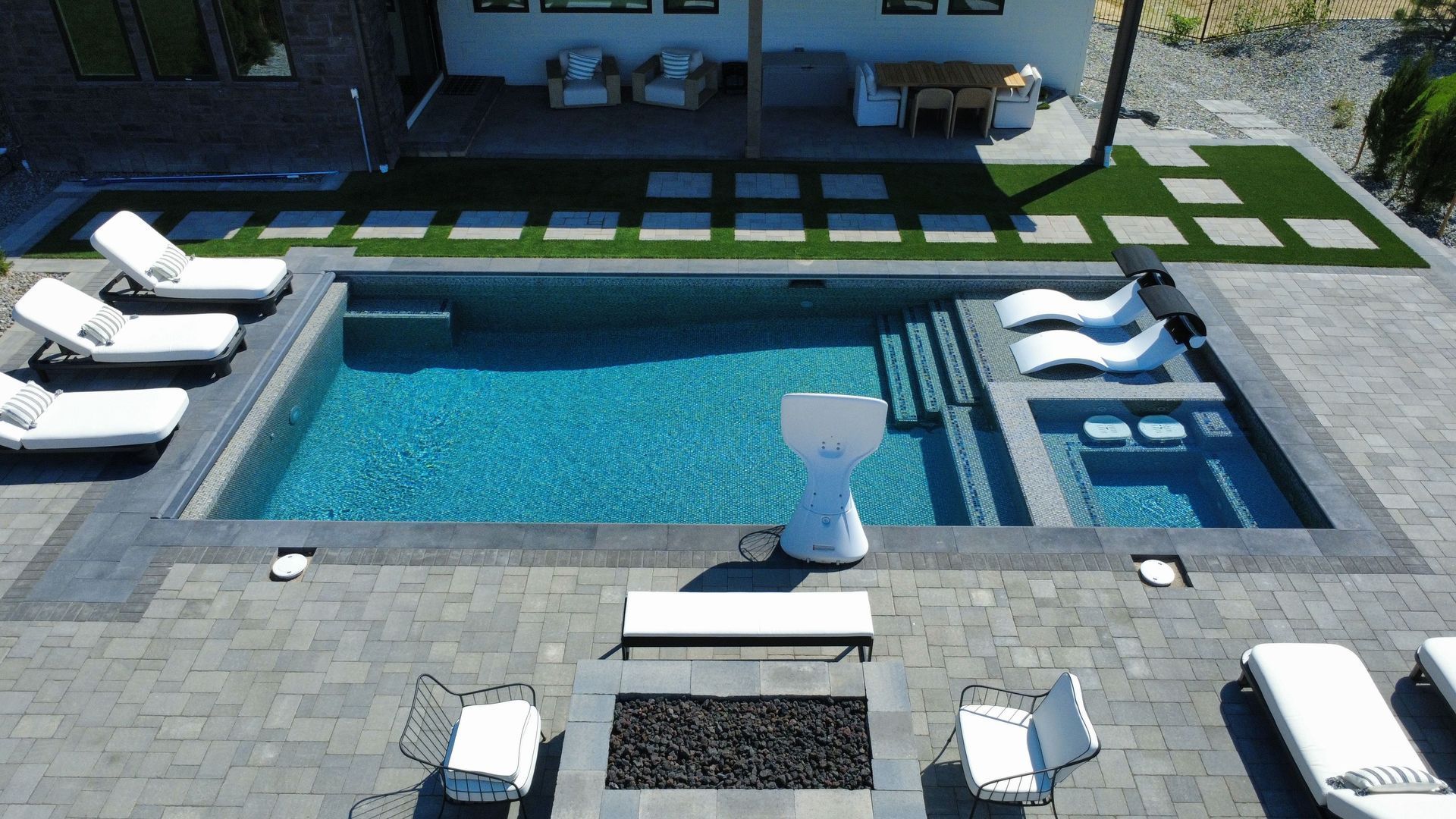Backyard pool with lounge chairs, steps, and a hot tub; surrounded by patio, turf, and a modern house.
