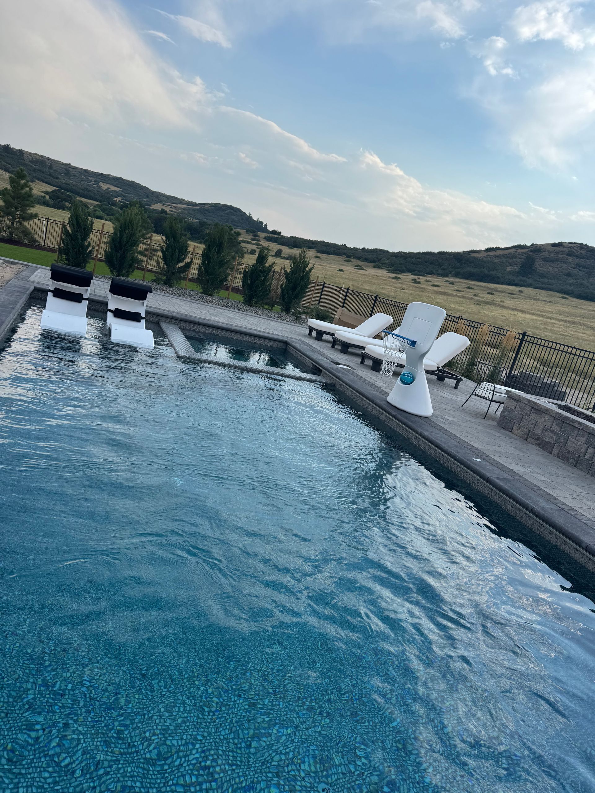 Infinity pool with blue water, overlooking a grassy field and hills under a partly cloudy sky.