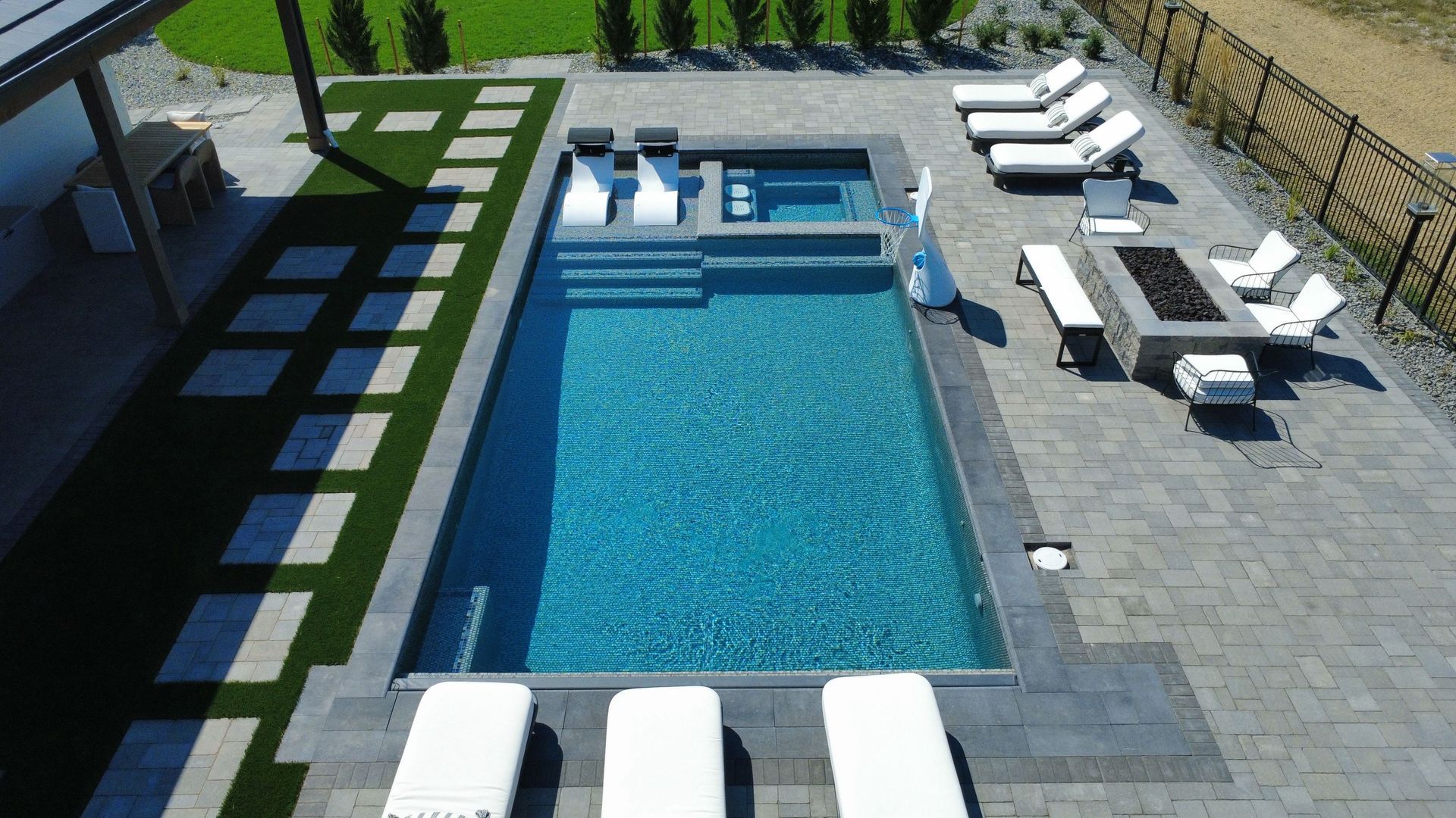Aerial view of a rectangular pool with a hot tub, lounge chairs, and a fire pit in a modern backyard.