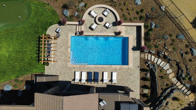 Aerial view of a rectangular swimming pool surrounded by a patio, seating area with a fire pit, and landscaping.