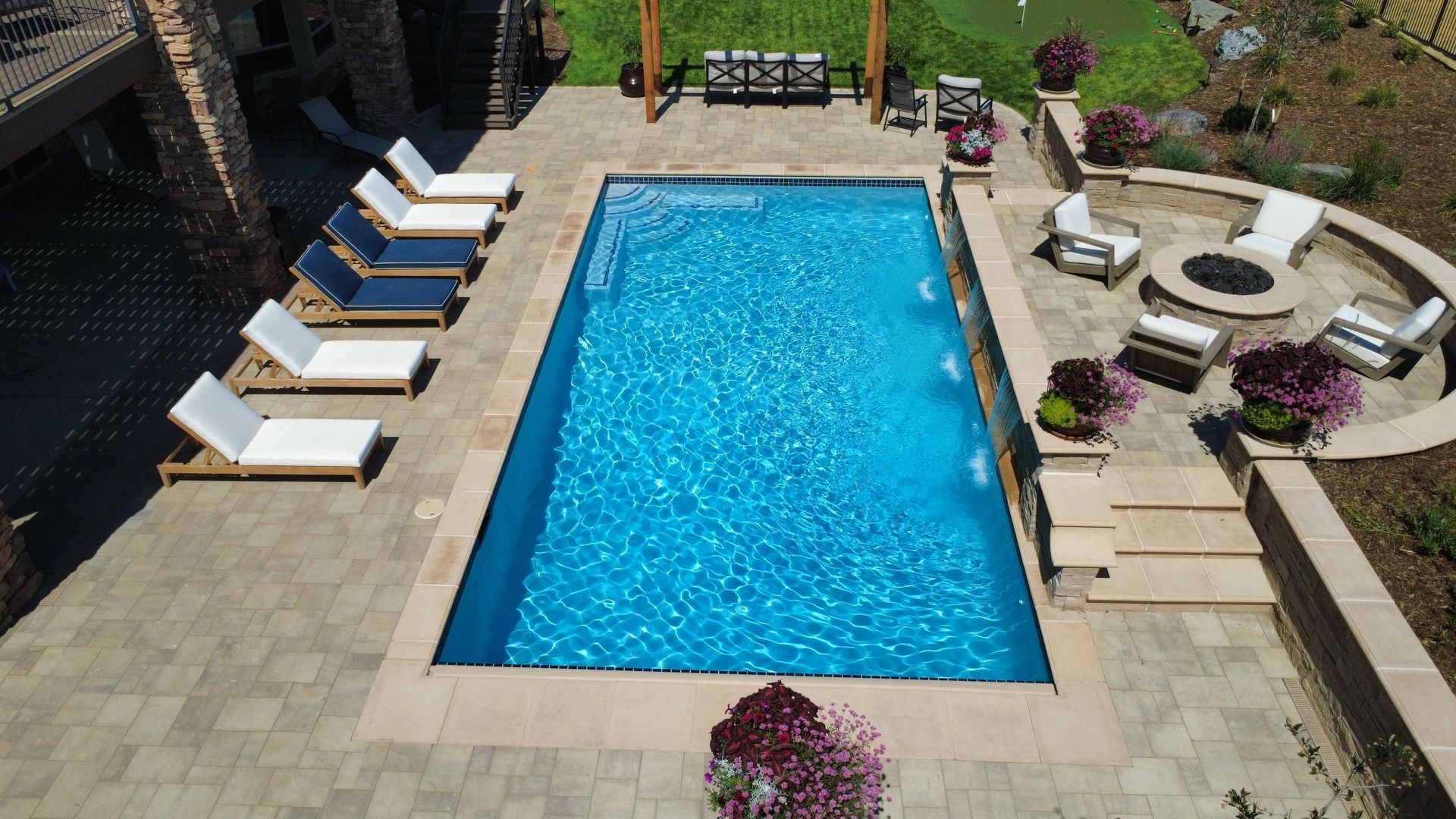 Rectangular pool in a stone patio, with lounge chairs, a fire pit area, and surrounding landscaping.