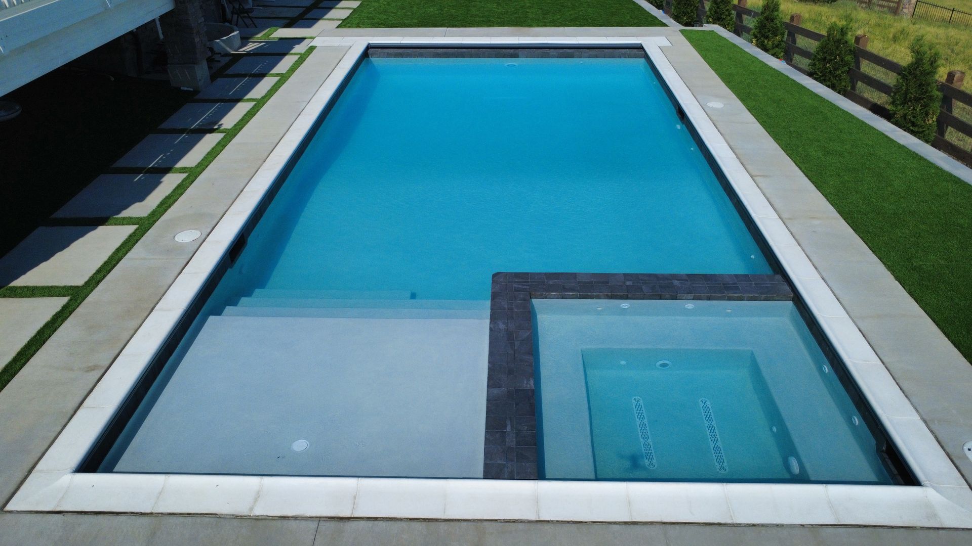 Rectangular pool with integrated steps, adjacent hot tub, light-colored stone surround, green grass, and fence.