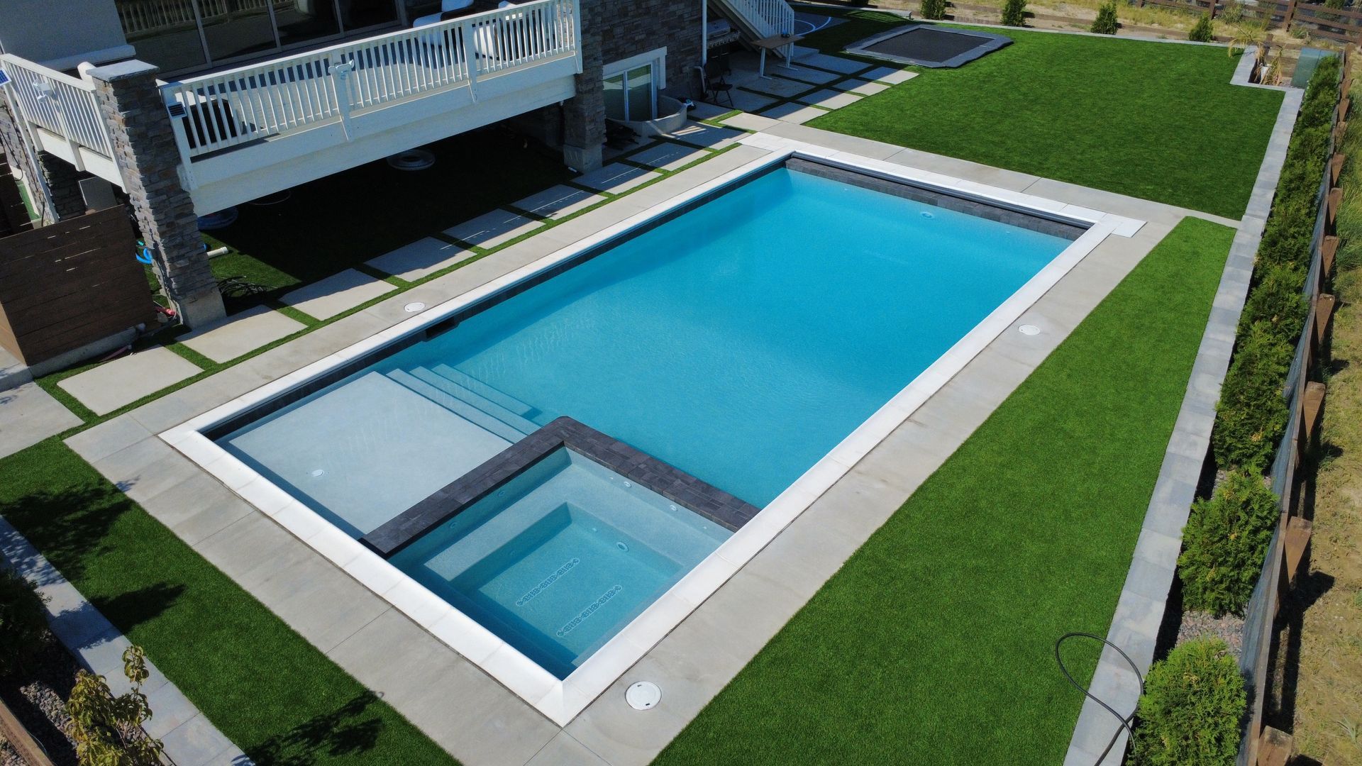Rectangular swimming pool with steps, surrounded by green turf and concrete patio, next to a white house.