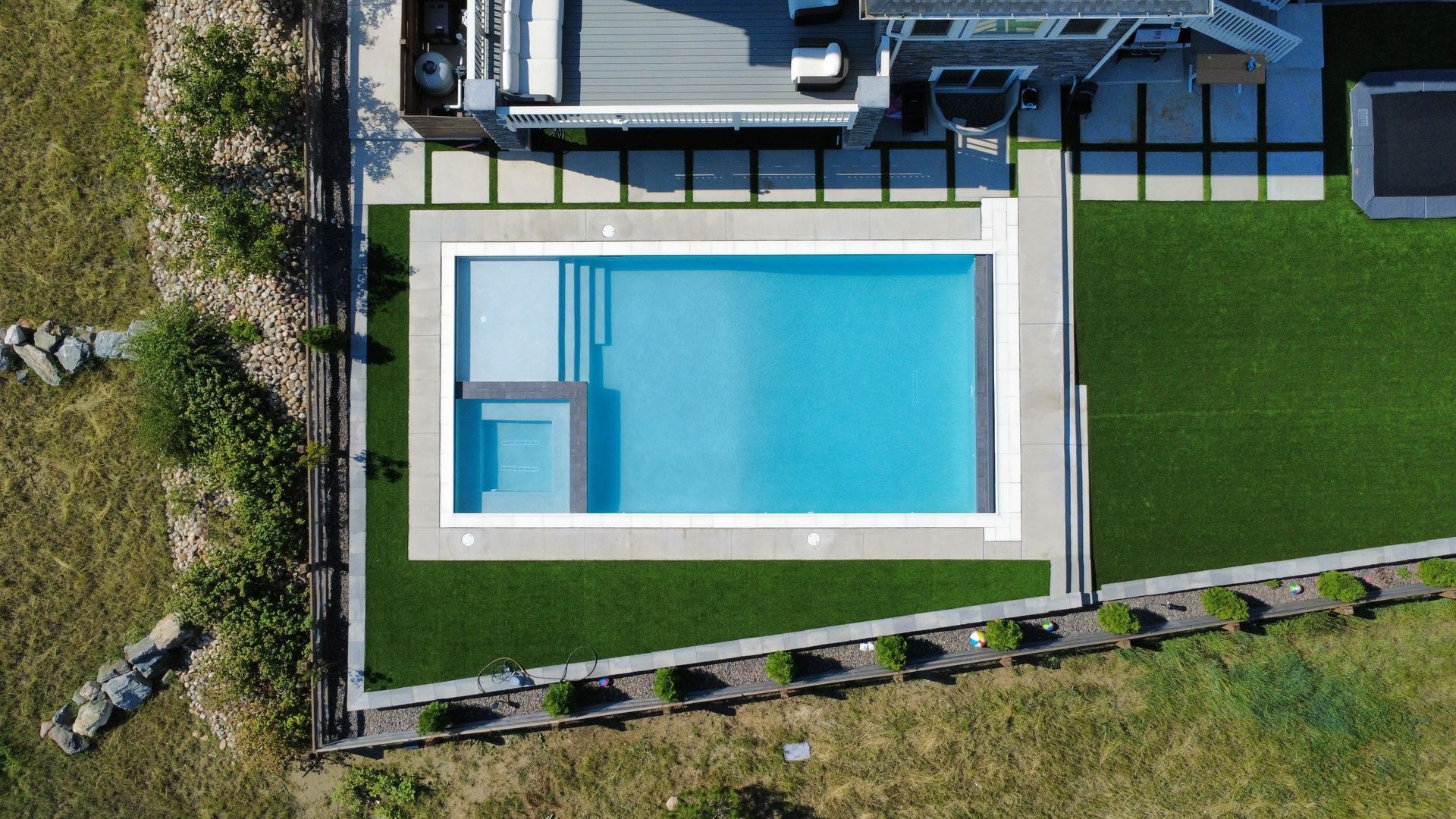 Aerial view of a rectangular pool with a spa, surrounded by concrete and artificial turf lawn.