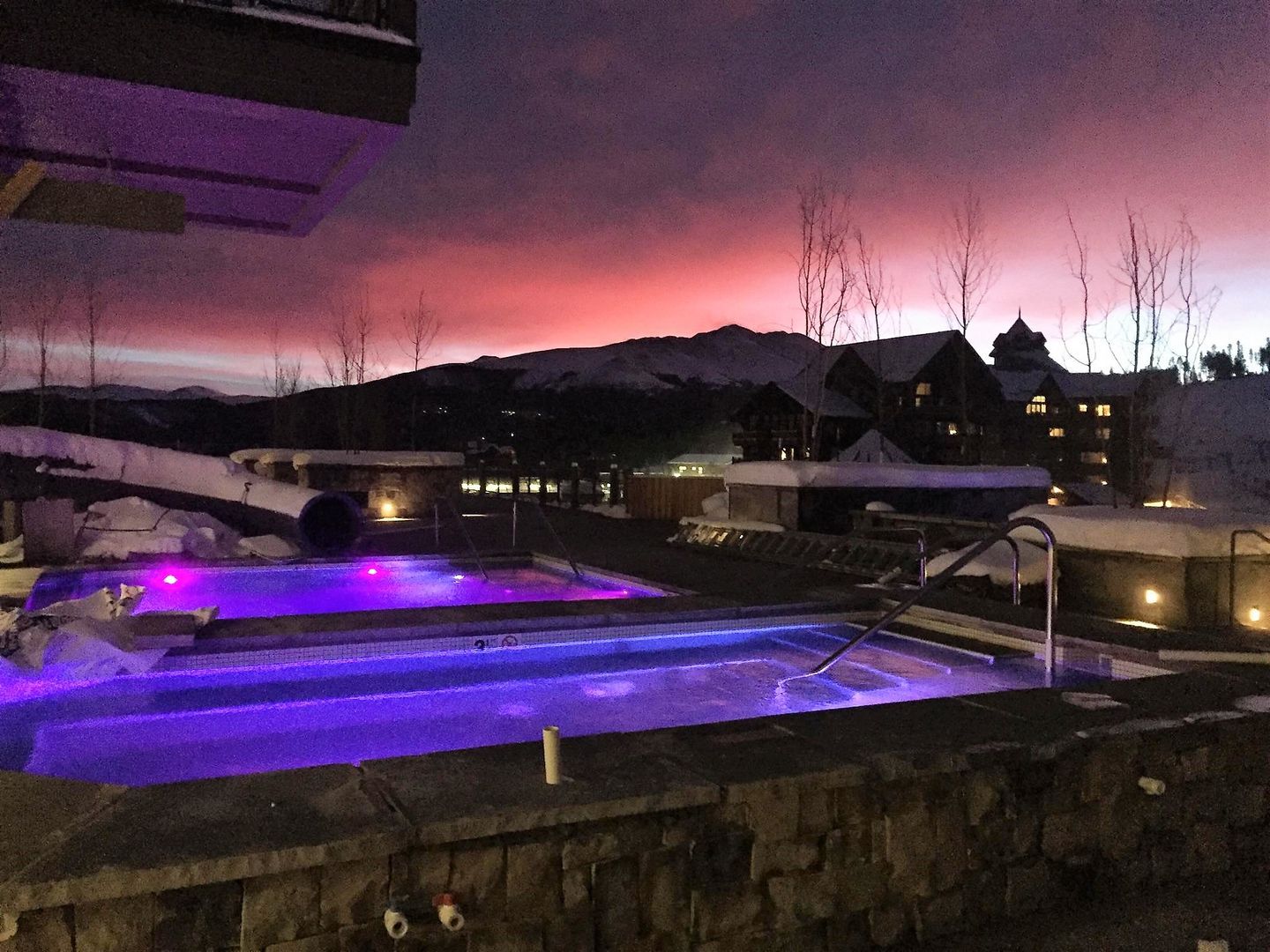 A swimming pool is lit up at night with a sunset in the background.