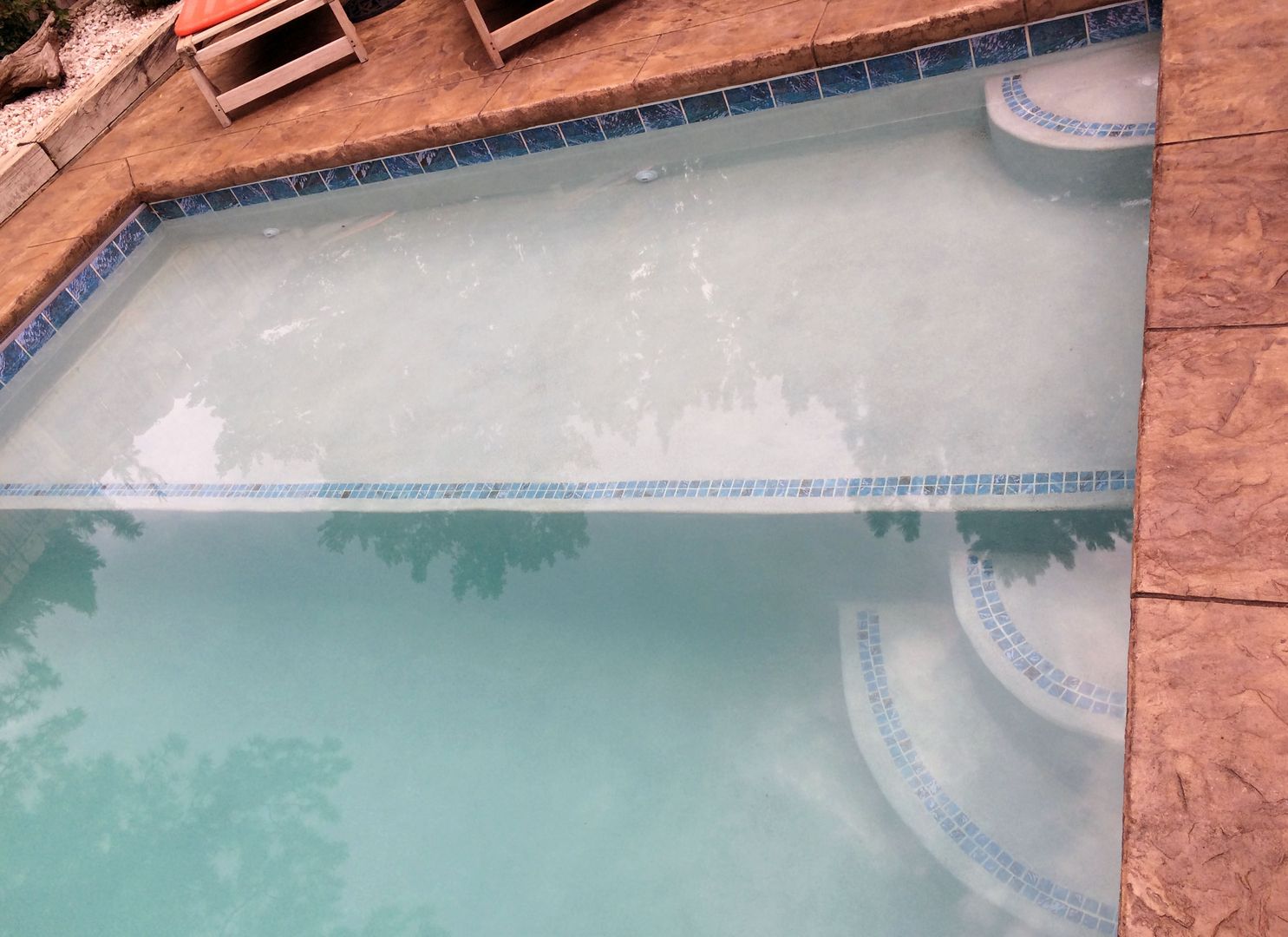 A swimming pool with stairs and a reflection of trees in the water.