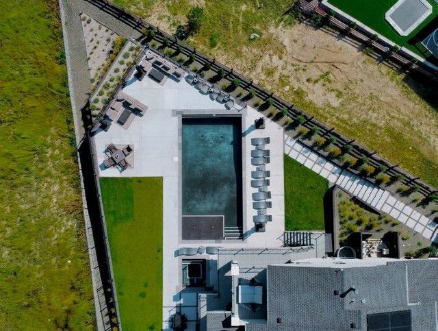 Aerial view of a rectangular pool surrounded by lounge chairs, with green grass and a paved area nearby.
