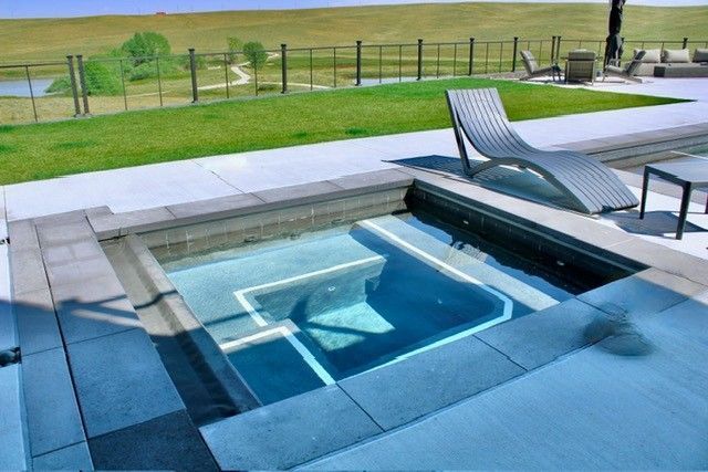 An outdoor hot tub with blue water, next to a pool with lounge chairs and a grassy landscape.