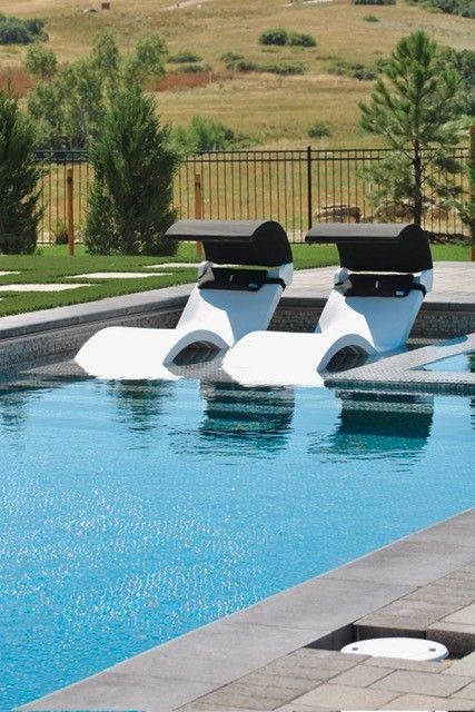 Two white, futuristic pool loungers with black canopies in a blue pool. Green lawn, trees, and a hill in background.
