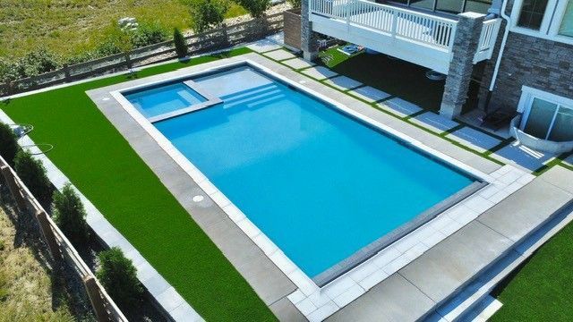 Rectangular pool with attached jacuzzi, bordered by green turf and concrete patio, next to a house.