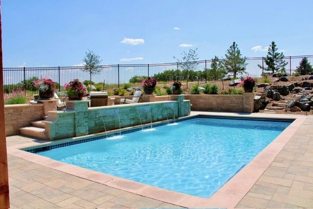 Backyard pool with a water feature, flower pots, and a grassy hill under a blue sky.