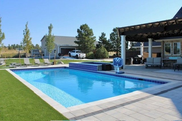Rectangular pool with blue water, surrounded by green grass, patio, and a house on a sunny day.