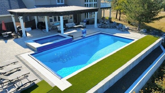 Backyard with a rectangular pool, spa, and seating area under a pergola. Green turf lines the pool's edge.