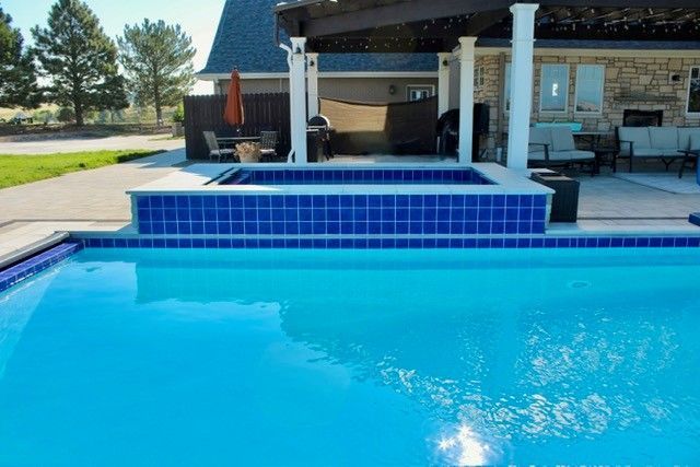 Blue pool with blue-tiled hot tub, patio, and house in the background. Bright sunny day.