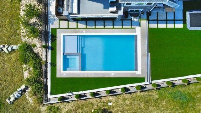 Aerial view of a blue rectangular pool with steps and a light-gray concrete border, surrounded by green turf.