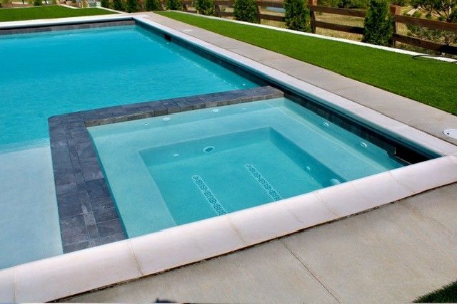 Swimming pool with a built-in spa; blue water, gray and white stone surround, green grass.
