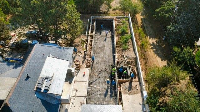 Construction of a long, rectangular concrete structure. Workers in blue and green work on the foundation. Green trees surround the site.