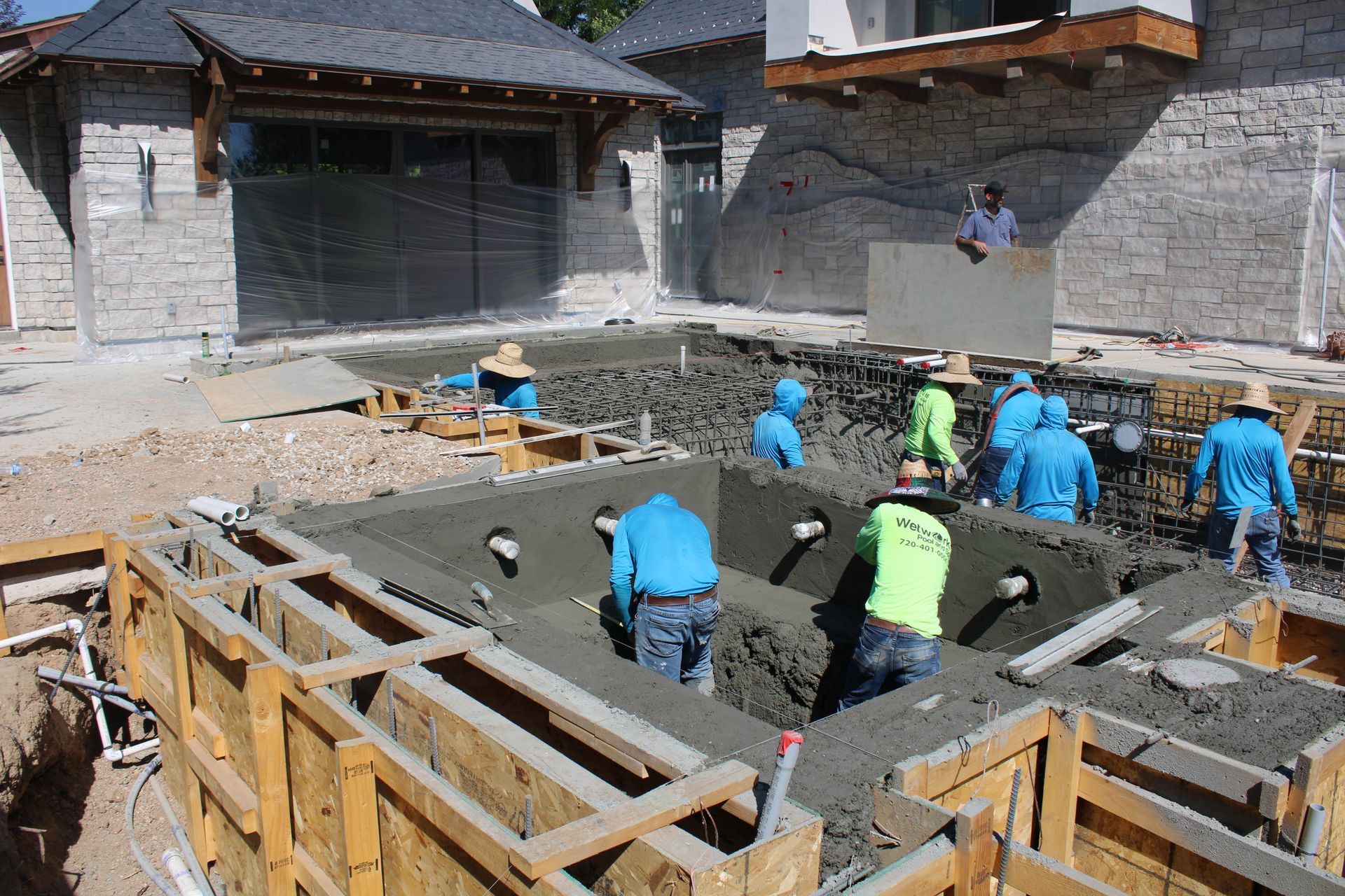 Construction workers pouring concrete into a pool form.  They are using shovels and wear blue and green shirts, hard hats.
