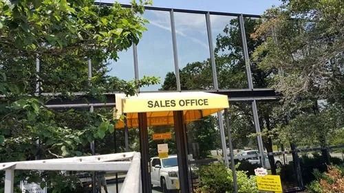 Sales office with yellow awning, glass windows, and trees. A white van is parked in front.
