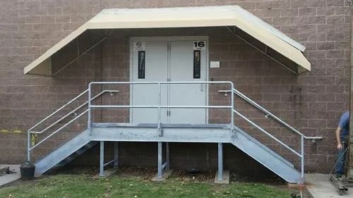 Metal steps leading to a building's double doors, protected by an awning. Handrails and a grassy area are visible.