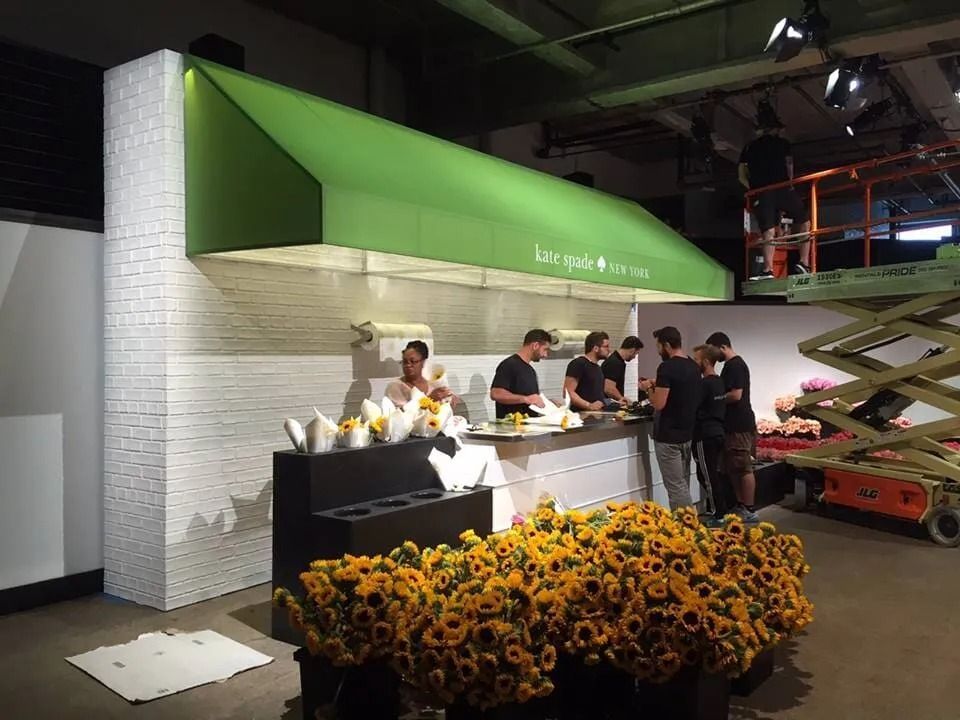 A flower shop with green awning and white brick facade. People are arranging flowers, sunflowers in the foreground.