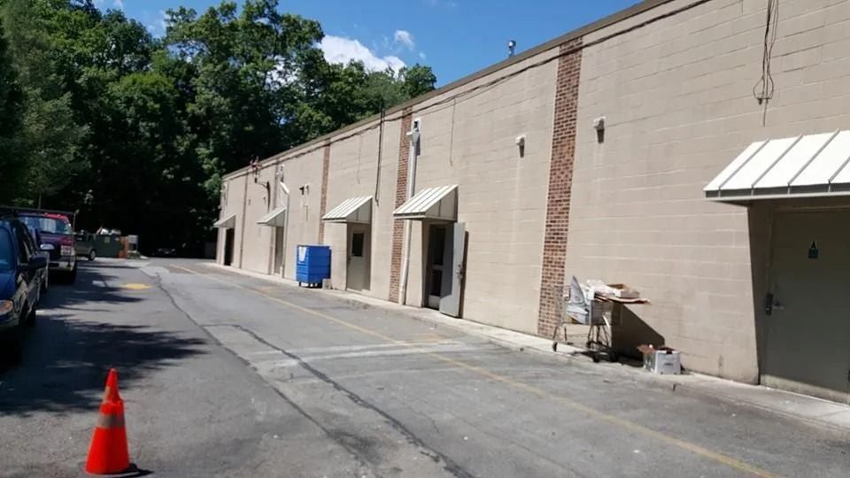 Long beige building with several doorways, awnings, and an asphalt road alongside.