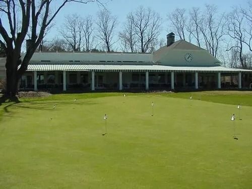 Green golf putting green in front of a white building with a covered porch and clock.