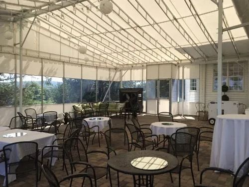 An open-air event space with round tables and chairs, a white canopy, and windows overlooking greenery.
