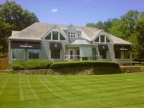 Gray building with awnings, set on a manicured lawn, stone wall, and trees in the background.
