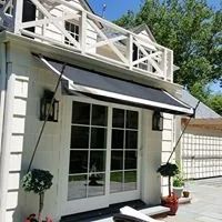 White house with black awning and balcony, trees in background.