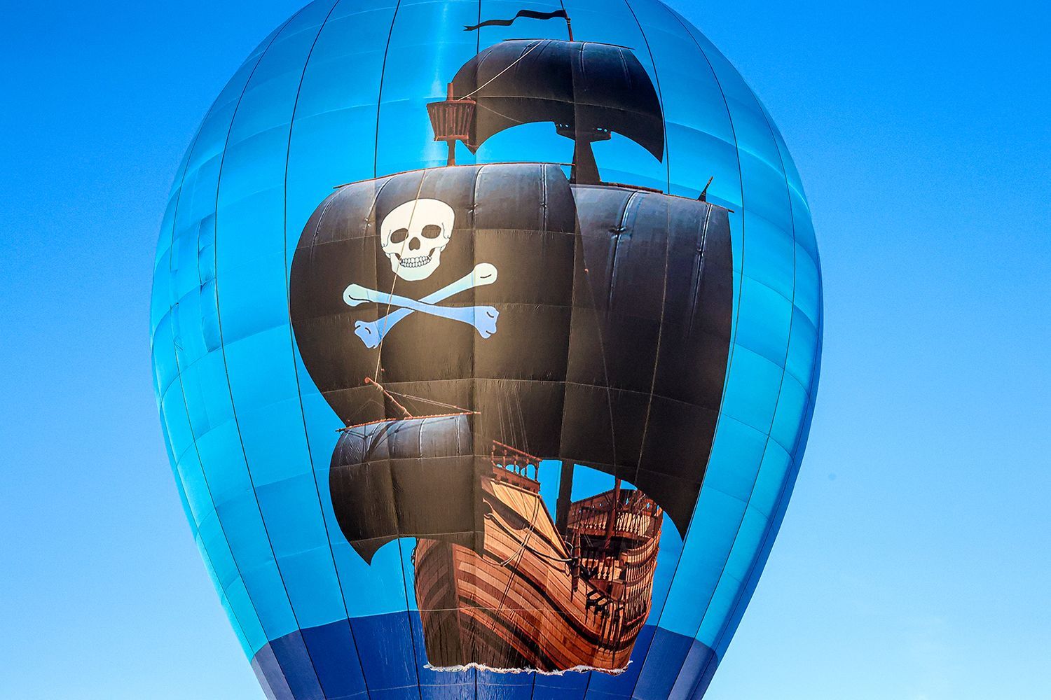 Blue hot air balloon shaped like a pirate ship with a skull and crossbones on a black sail, against a clear blue sky.