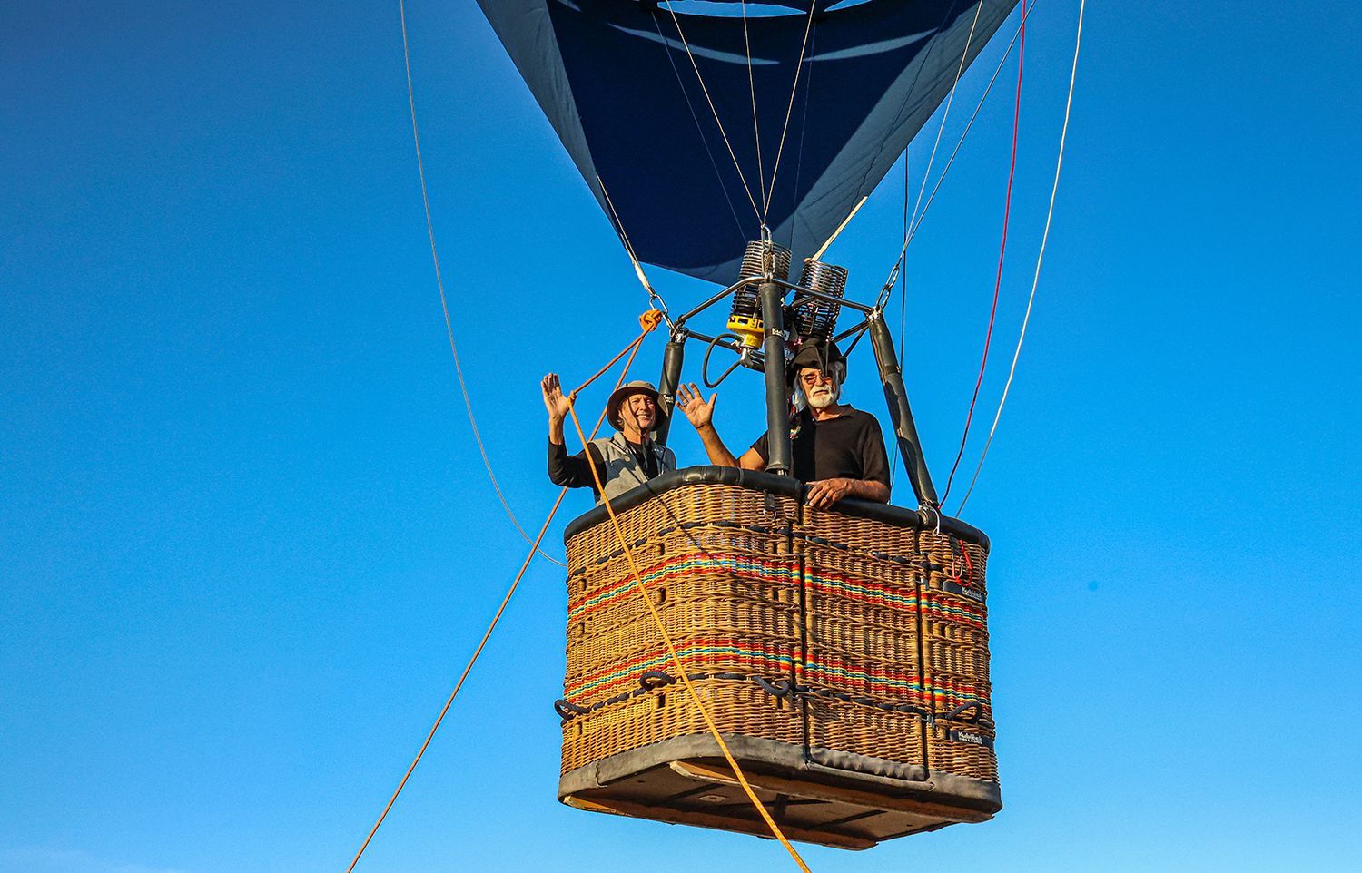 Two people waving from a hot air balloon basket against a clear blue sky.